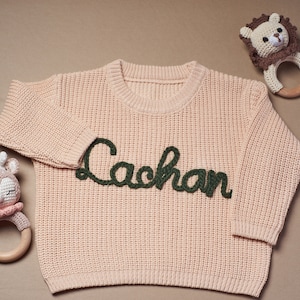 May include: A light pink baby sweater with the name "Lachan" embroidered in dark green. The sweater is surrounded by two crochet baby rattles, one shaped like a lion and the other like a deer.