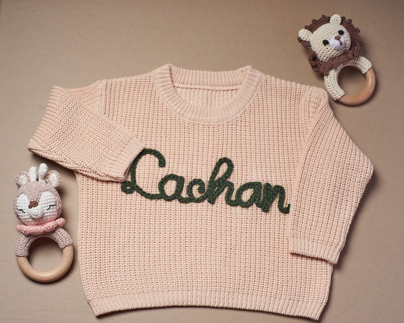 May include: A light pink baby sweater with the name "Lachan" embroidered in dark green. The sweater is surrounded by two crochet baby rattles, one shaped like a lion and the other like a deer.