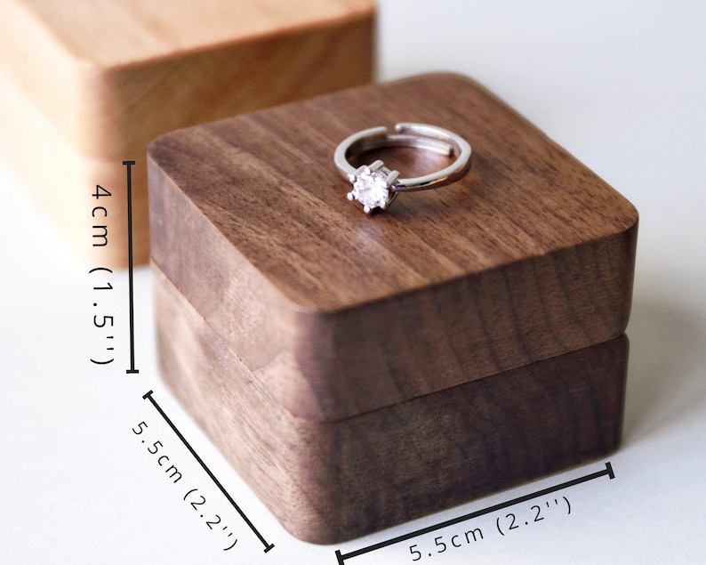 Op de afbeelding: Een vierkante houten ringdoos met een donkerbruine afwerking. De doos is 5,5 cm breed en 4 cm hoog. Een zilveren ring met een heldere steen wordt op de bovenkant van de doos getoond.