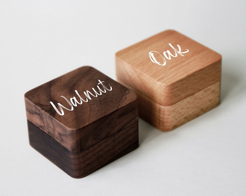 Op de afbeelding: Twee houten ringendoosjes, een van walnoot en een van eikenhout. De doosjes zijn vierkant en hebben een scharnierend deksel. De woorden "Walnoot" en "Eikenhout" zijn gegraveerd op de bovenkant van elk doosje.