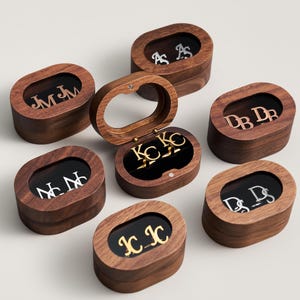 May include: An assortment of wooden jewelry boxes with oval-shaped lids. Each box contains initials in various metallic finishes, including gold and silver. The boxes are arranged on a light background, with one box open, revealing its contents.