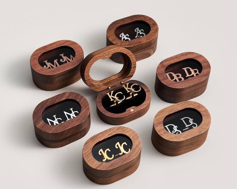 May include: An assortment of wooden jewelry boxes with oval-shaped lids. Each box contains initials in various metallic finishes, including gold and silver. The boxes are arranged on a light background, with one box open, revealing its contents.