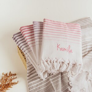 Op de afbeelding: Een stapel gestreepte handdoeken met een roze en witte handdoek bovenop. De roze en witte handdoek heeft de naam "Kamila" geborduurd in roze draad.