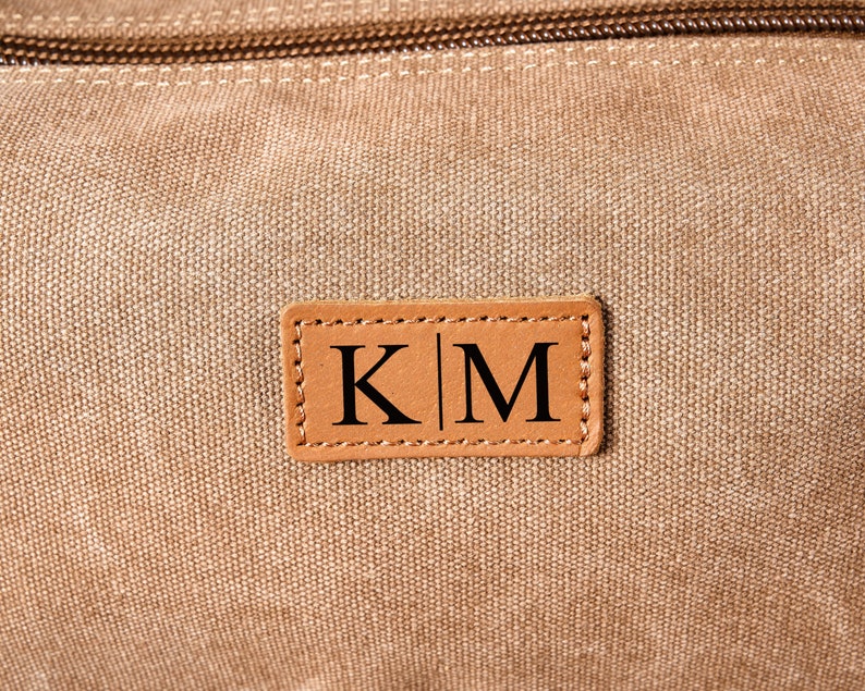 Puede incluir: Una bolsa de lona marr&oacute;n con un parche de cuero que dice "K|M" en letras negras.