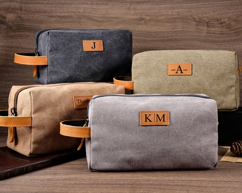 Puede incluir: Cuatro neceseres de lona con asas de cuero y parches de cuero personalizados. Las bolsas son de color gris, marr&oacute;n, verde y negro. Los parches tienen las iniciales "J", "A", "KM" y "Thom".