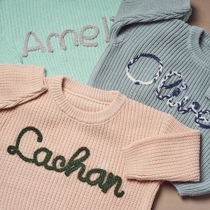 May include: Three knitted sweaters in pastel colors: mint green, dusty rose, and blue-gray. Each sweater has a name embroidered on the front in a contrasting color. The names are "Amelia", "Lachlan", and "Oliver".