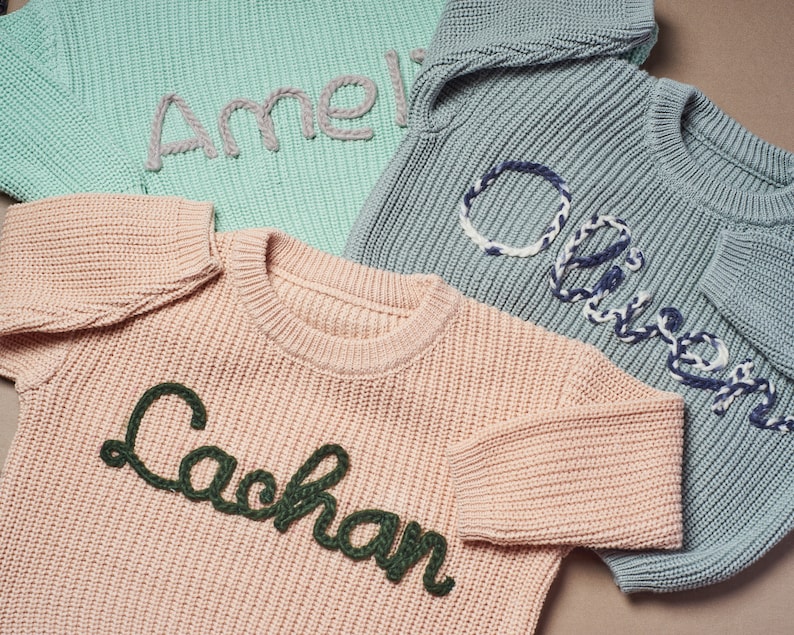 May include: Three knitted sweaters in pastel colors: mint green, dusty rose, and blue-gray. Each sweater has a name embroidered on the front in a contrasting color. The names are "Amelia", "Lachlan", and "Oliver".