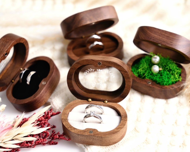 May include: Three wooden ring boxes with different interiors. One box has a black velvet interior, another has a white velvet interior, and the third has a green moss interior. The box with the white velvet interior has a clear glass lid with the words "Mr. & Mrs." engraved on it.