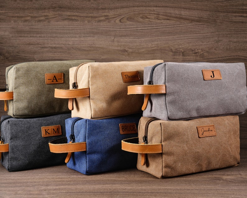 Puede incluir: Seis bolsas de aseo de lona con asas de cuero e iniciales personalizadas. Las bolsas est&aacute;n apiladas en forma de pir&aacute;mide sobre una superficie de madera. Las iniciales son "A", "Thomas", "J", "KM", "Bruce" y "Jacob".