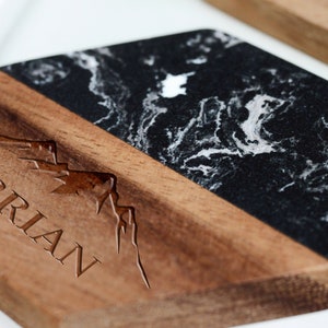 May include: A hexagonal coaster with a black and white marble top and a wooden base. The base has a mountain range design and the name "BRIAN" engraved in the wood.