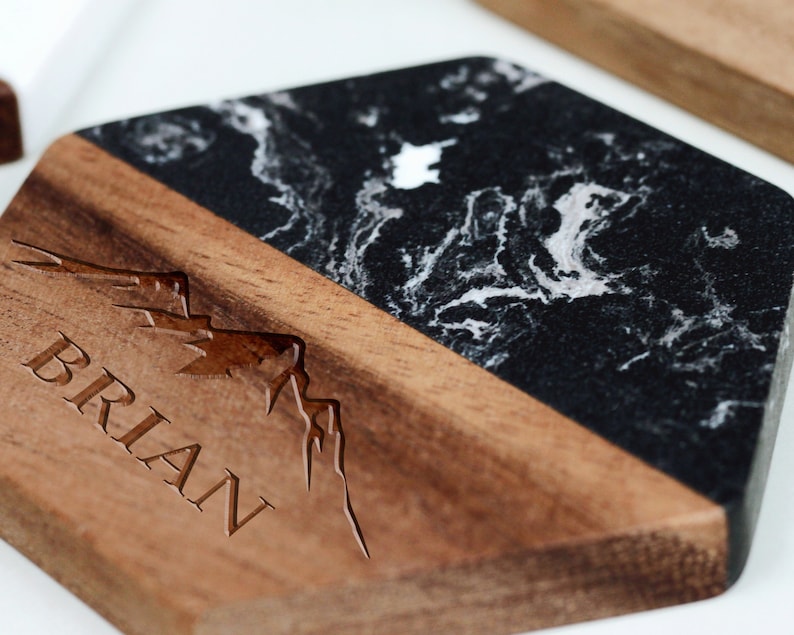 May include: A hexagonal coaster with a black and white marble top and a wooden base. The base has a mountain range design and the name "BRIAN" engraved in the wood.