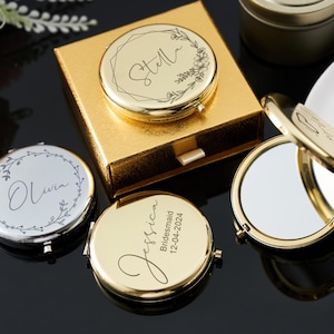 May include: Gold and silver compact mirrors with personalised text and floral designs. One mirror is open, revealing a reflective surface. The mirrors are on a reflective black surface, with a gold box in the background.