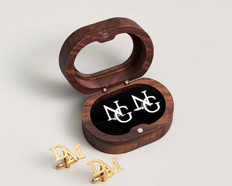 May include: A wooden oval-shaped box with a clear top, containing white cufflinks with the letters "NG". Two gold cufflinks with the letters "DM" are displayed below the box. The box is open, revealing a black interior.