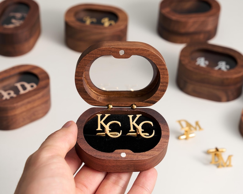 May include: A wooden oval-shaped box with a clear top, open to reveal gold-colored cufflinks with the initials "KC". Other similar boxes with different initials are visible in the background. The boxes are made of dark wood.