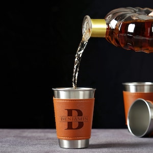 Peut inclure: Verres à shot en acier inoxydable avec des manchons en cuir marron. Les manchons en cuir sont gravés de la lettre "B" et du nom "Benjamin". Une bouteille de liquide ambré est versée dans l'un des verres.