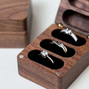May include: A wooden ring box with three compartments, each lined with black velvet. The box is made of dark wood and has a polished finish. Three silver rings with diamonds are displayed in the compartments.