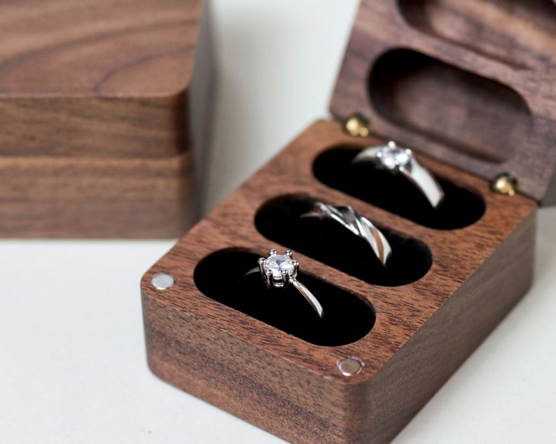 May include: A wooden ring box with three compartments, each lined with black velvet. The box is made of dark wood and has a polished finish. Three silver rings with diamonds are displayed in the compartments.