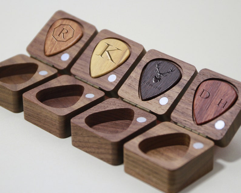 May include: A wooden box with six compartments, each containing a personalized guitar pick. The picks are made of different materials and feature various designs, including initials, a deer, and a geometric pattern.
