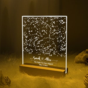 Puede incluir: Una luz LED blanca con una base de madera muestra un mapa de constelaciones con los nombres "Sarah & Alix" y el texto "The Night Our Stars Aligned May 15 2014".