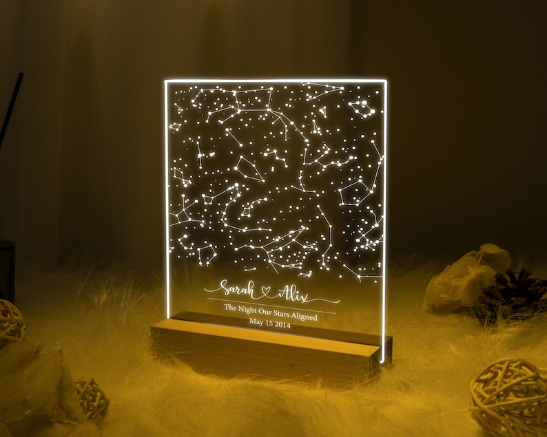 Puede incluir: Una luz LED blanca con una base de madera muestra un mapa de constelaciones con los nombres "Sarah & Alix" y el texto "The Night Our Stars Aligned May 15 2014".