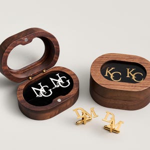May include: Wooden oval cufflink boxes with gold and silver initial cufflinks. One box is open, revealing silver initials on a black velvet interior. The other box is closed, with gold initials visible through a clear top. Two pairs of gold cufflinks are displayed.