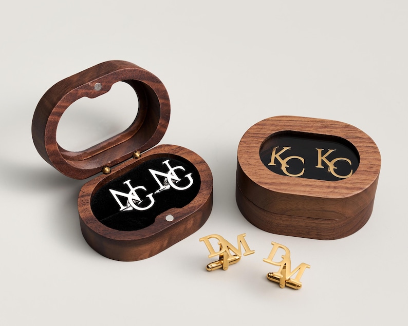 May include: Wooden oval cufflink boxes with gold and silver initial cufflinks. One box is open, revealing silver initials on a black velvet interior. The other box is closed, with gold initials visible through a clear top. Two pairs of gold cufflinks are displayed.