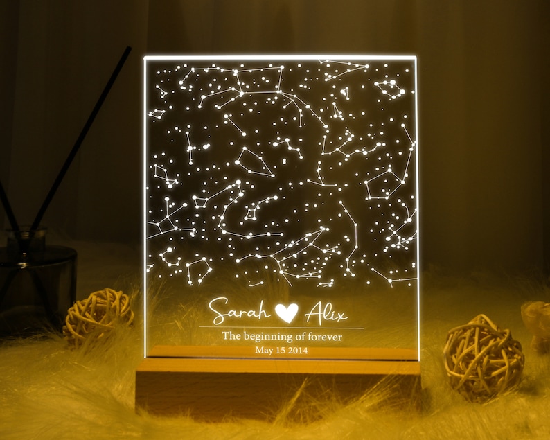 Puede incluir: Una luz LED blanca con un dise&ntilde;o de constelaci&oacute;n y los nombres "Sarah" y "Alix" con un s&iacute;mbolo de coraz&oacute;n entre ellos. El texto debajo dice "The beginning of forever" y "May 15 2014". La luz est&aacute; sobre una base de madera.