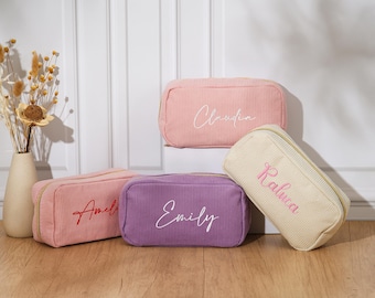 May include: A collection of personalized corduroy cosmetic bags in various colors: light pink, purple, and cream. Each bag features a name embroidered in a contrasting color. The bags have a rectangular shape with a zipper closure, perfect for storing makeup or toiletries.