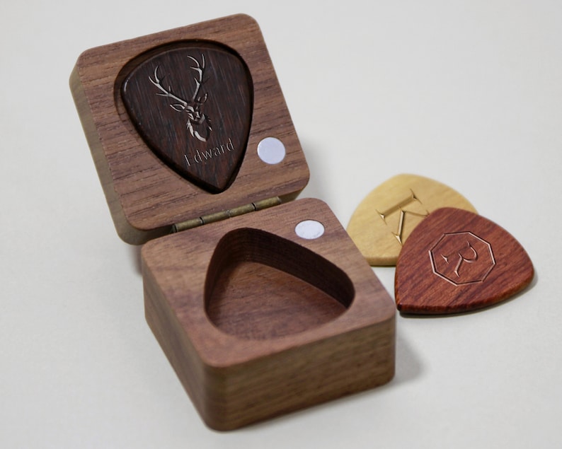 May include: A wooden box with a hinged lid, containing a dark brown guitar pick with a deer head design and the word "Edward" engraved on it. Three additional guitar picks are visible outside the box, one light brown with a letter "Z" and two dark red with a geometric design.