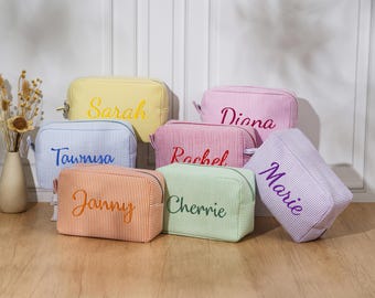 May include: A collection of personalized cosmetic bags in various colors, including yellow, blue, orange, green, pink, and purple. Each bag features a vertical striped pattern and a name embroidered in a contrasting color.