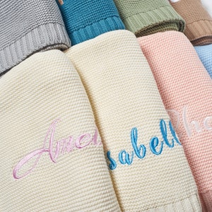 May include: A stack of knitted baby blankets in various colors, including gray, teal, green, beige, and light blue. The blankets are folded and have embroidered names on them, including "Amelia", "Isabelle", and "Lily".