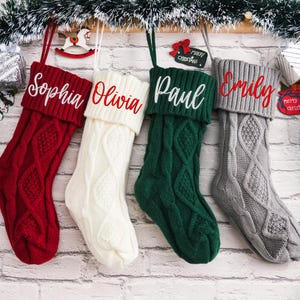 May include: Four knitted Christmas stockings with names embroidered on them. The stockings are red, white, green, and gray. The names are Sophia, Olivia, Paul, and Emily.