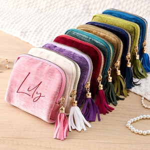 Puede incluir: Una colección de neceseres de terciopelo de colores, con cremalleras doradas y borlas a juego. La bolsa rosa tiene el nombre "Lily" impreso. Los neceseres están colocados sobre una superficie de madera.