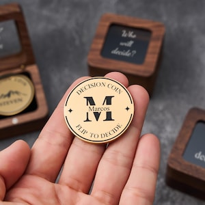 May include: A gold-colored decision coin with the text "DECISION COIN" and "FLIP TO DECIDE". The coin features the name "Marcos" and the letter "M". The coin is held in a hand, with wooden boxes in the background.