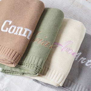 May include: Four personalized knitted baby blankets in different colors. The blankets are stacked on top of each other. The top blanket is brown with the name "Connor" embroidered in white. The second blanket is green with the name "Olivia" embroidered in white. The third blanket is white with the name "Amelia" embroidered in pink. The bottom blanket is gray with the name "Arthur" embroidered in white.