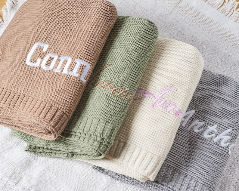 May include: Four personalized knitted baby blankets in different colors. The blankets are stacked on top of each other. The top blanket is brown with the name "Connor" embroidered in white. The second blanket is green with the name "Olivia" embroidered in white. The third blanket is white with the name "Amelia" embroidered in pink. The bottom blanket is gray with the name "Arthur" embroidered in white.