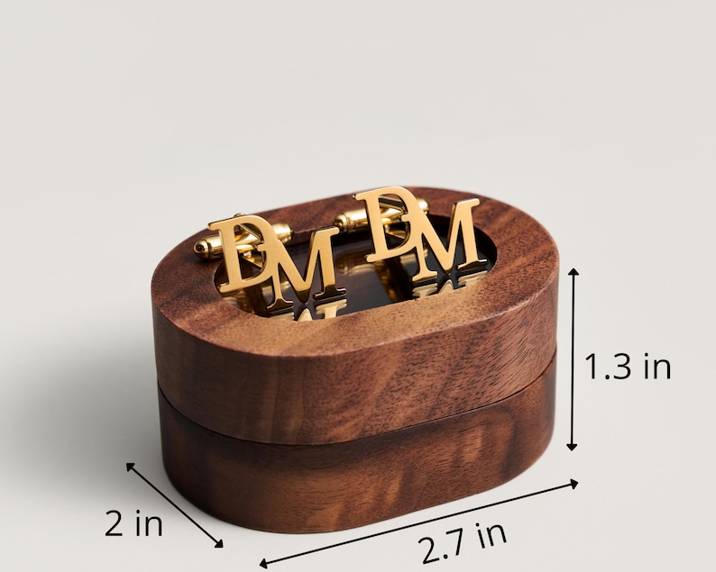 May include: A wooden oval box with gold-colored cufflinks. The cufflinks feature the letters "DM". The box is 2.7 inches long, 2 inches wide, and 1.3 inches tall. The box and cufflinks are on a light background.