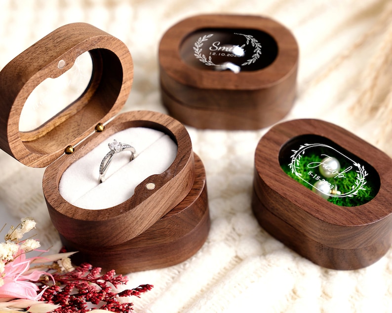 May include: Three wooden ring boxes with clear glass lids. The first box has a ring inside, the second box has a floral design and the text "Smith 12.10.2023", and the third box has a floral design and the text "Jess 8.18.2023".