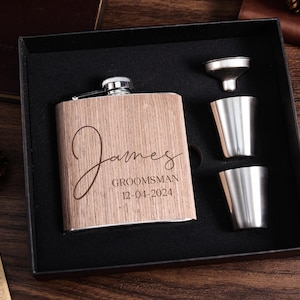 May include: A wooden flask with the name "James" engraved on it, along with the text "Groomsman 12-04-2024". The flask is in a black box with two shot glasses and a funnel.