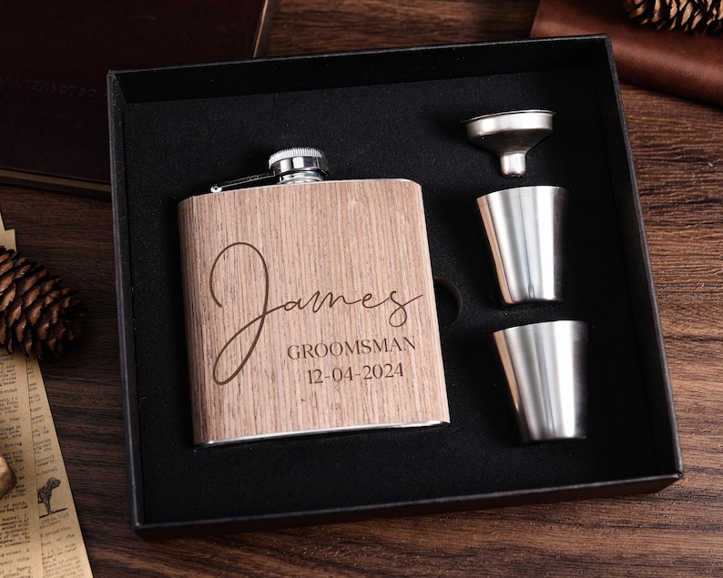 May include: A wooden flask with the name "James" engraved on it, along with the text "Groomsman 12-04-2024". The flask is in a black box with two shot glasses and a funnel.