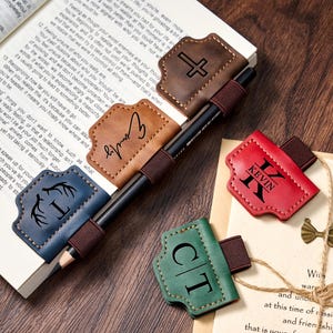 Può includere: Set di segnalibri in pelle in vari colori, tra cui marrone, blu, rosso e verde. Ogni segnalibro presenta un design unico, come una croce, delle iniziali o un nome. Una penna è inserita nell'elastico dei segnalibri.