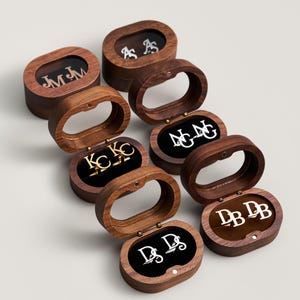May include: A collection of personalized cufflinks and earring sets displayed in open wooden oval boxes. Each set features initials in silver or gold against a black velvet interior. The boxes are made of dark wood.
