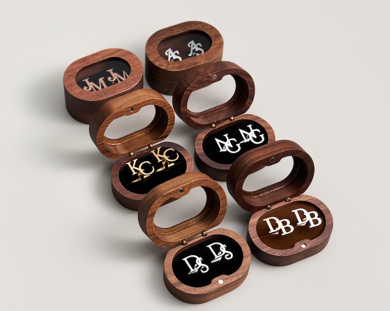 May include: A collection of personalized cufflinks and earring sets displayed in open wooden oval boxes. Each set features initials in silver or gold against a black velvet interior. The boxes are made of dark wood.