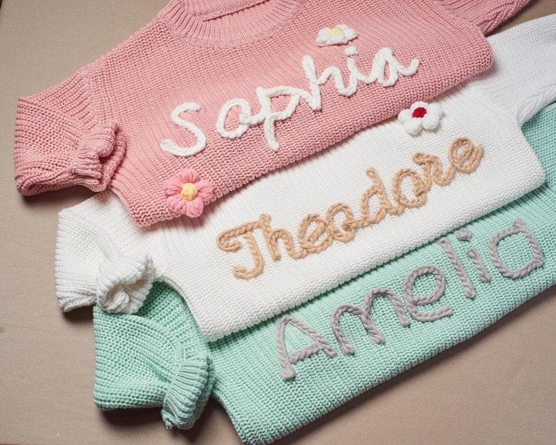 May include: Close-up of three pastel-colored knit sweaters. The pink sweater has the name "Sophia" in white yarn and a flower applique. The white sweater says "Theodore" in tan yarn. The green sweater says "Amelia" in gray yarn. All sweaters have ribbed cuffs.