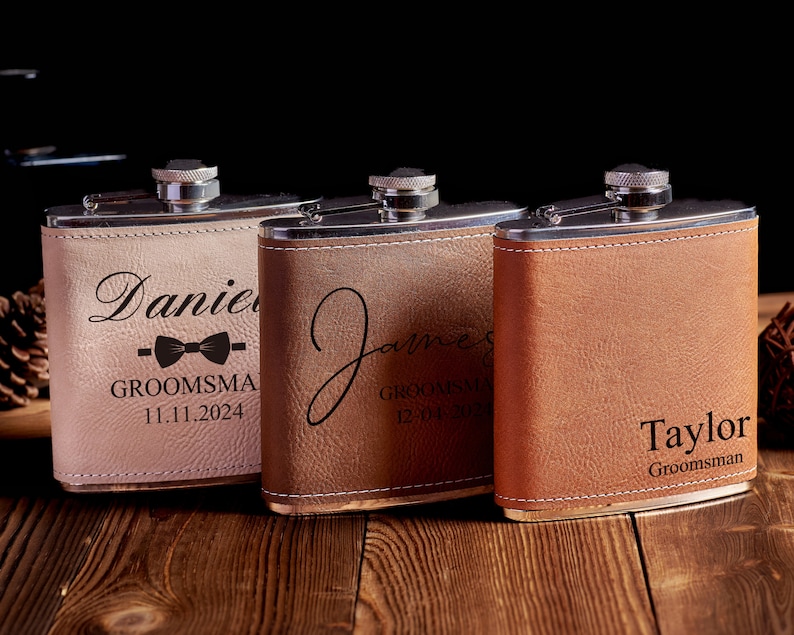 May include: Three personalized stainless steel hip flasks with leather coverings. The flasks are engraved with the names "Daniel", "James", and "Taylor" along with the title "Groomsman" and a date.