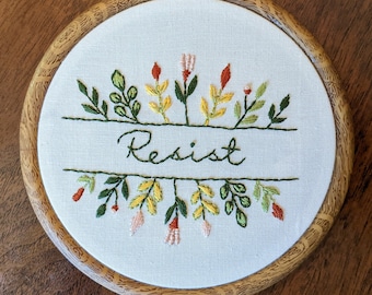 May include: Embroidered hoop art with a white background and a wooden frame. The embroidery features green, yellow, and red floral designs with the word "Resist" stitched in cursive.