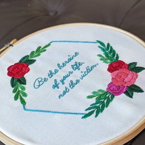 May include: Embroidered hoop art featuring a teal blue frame and the quote "Be the heroine of your life, not the victim." The quote is surrounded by green leaves and red and pink roses.