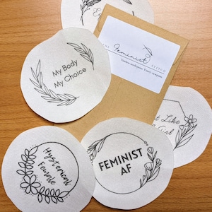 May include: Five white fabric patches with black ink designs and text. The patches feature feminist slogans, including "My Body My Choice", "Hysterical Female", "Feminist AF", and "Don't Like a Girl".