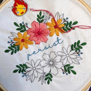May include: A white embroidery hoop with a floral design and the word "persist" stitched in blue thread. The hoop is surrounded by a wooden frame.