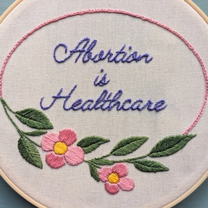 May include: Embroidered hoop art with a pink floral design and the text "Abortion is Healthcare" in purple thread.